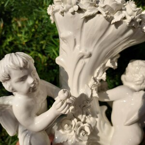 May include: White porcelain vase with two cherubs, one holding an apple, and a floral design around the top.