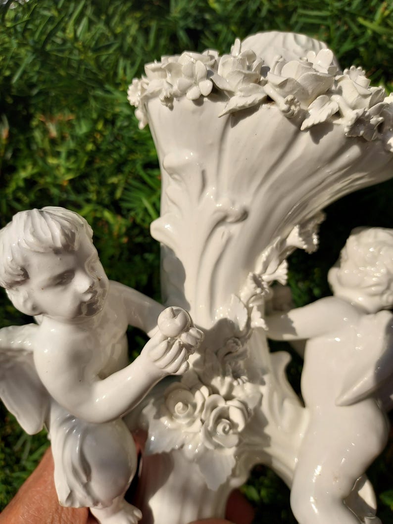 May include: White porcelain vase with two cherubs, one holding an apple, and a floral design around the top.