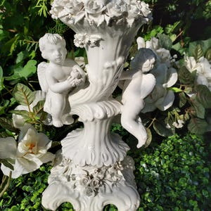 May include: White porcelain candle holder with two cherubs and floral accents. The holder is decorated with roses and has a scrolled design.