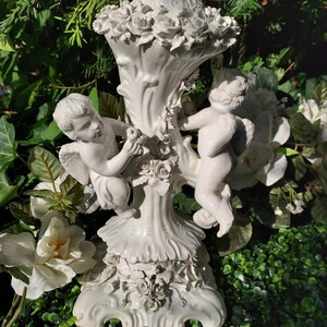 May include: White ceramic candle holder with two cherubs holding a flower. The holder is decorated with roses and has a scalloped base.