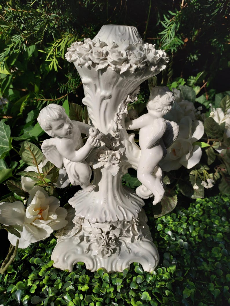 May include: White ceramic candle holder with two cherubs holding a flower. The holder is decorated with roses and has a scalloped base.