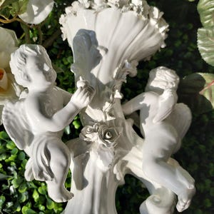 May include: White porcelain vase with two cherubs holding roses. The vase is decorated with floral details and has a scalloped top.