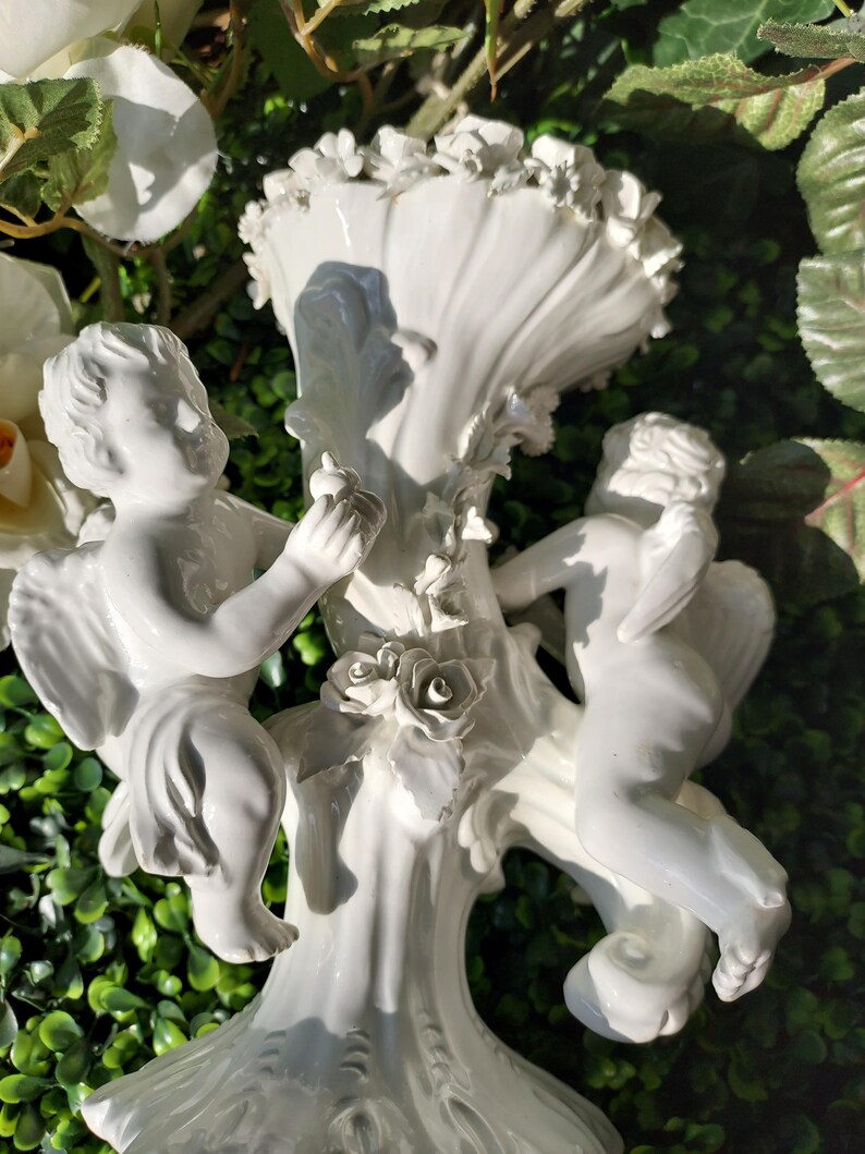 May include: White porcelain vase with two cherubs holding roses. The vase is decorated with floral details and has a scalloped top.