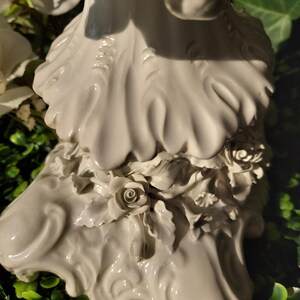 May include: White porcelain figurine of a woman with a flowing gown and floral details. The base of the figurine is decorated with sculpted roses and leaves.