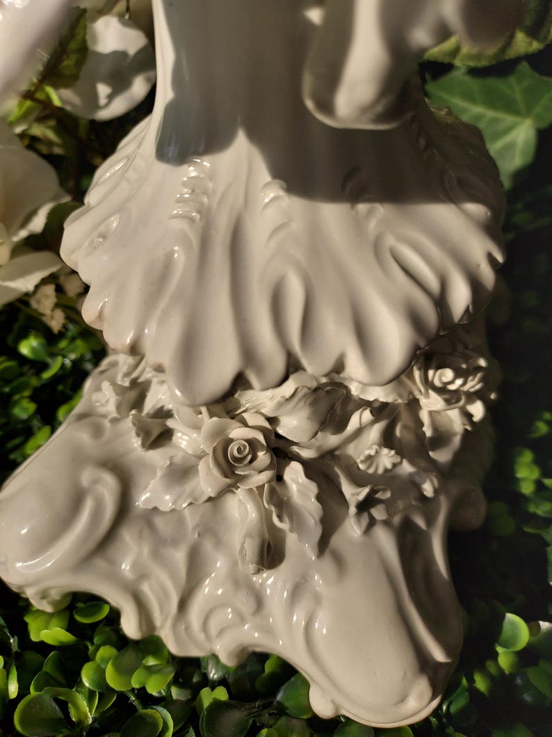 May include: White porcelain figurine of a woman with a flowing gown and floral details. The base of the figurine is decorated with sculpted roses and leaves.