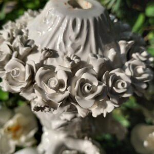 May include: White porcelain figurine of a cherub holding a bouquet of roses. The cherub is sitting on a base with a floral design. The figurine is a decorative piece for the home.