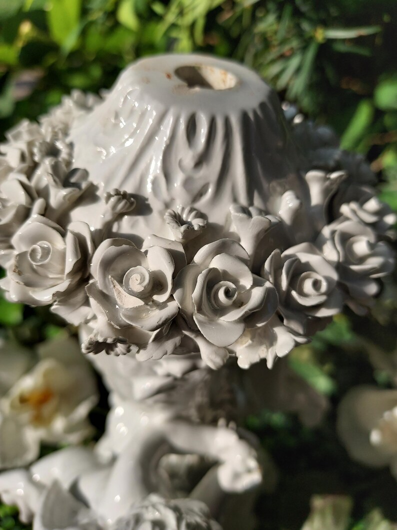 May include: White porcelain figurine of a cherub holding a bouquet of roses. The cherub is sitting on a base with a floral design. The figurine is a decorative piece for the home.