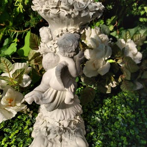 May include: White ceramic candle holder featuring two cherubs holding a floral design. The base is decorated with roses and intricate details.
