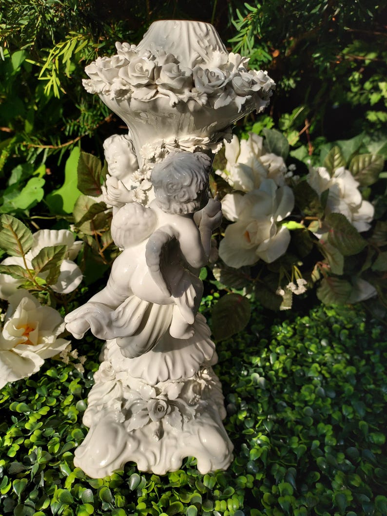 May include: White ceramic candle holder featuring two cherubs holding a floral design. The base is decorated with roses and intricate details.