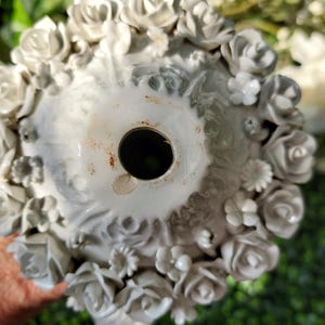 May include: White ceramic vase with a floral design. The vase is decorated with raised roses and other flowers. The vase has a hole in the bottom for drainage.