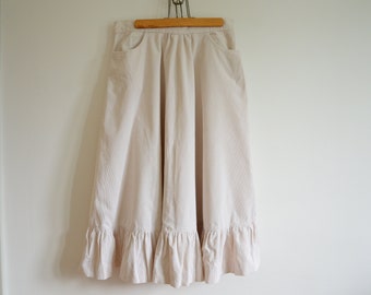 May include: A white corduroy skirt with a ruffled hem. The skirt has two pockets and is hanging on a wooden hanger.