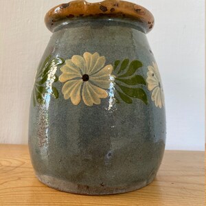 May include: A blue-green ceramic vase with a wide, slightly irregular rim. The vase is decorated with a band of cream-coloured flowers with dark centres and green leaves. The rim has a brown, textured finish.