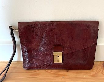May include: A burgundy leather clutch bag with a gold clasp and a black strap.