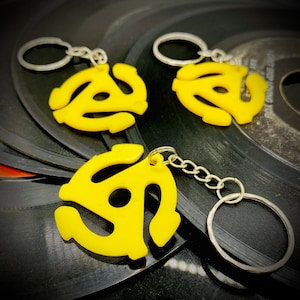 May include: Three yellow 45 RPM record adapter keychains with silver key rings. The keychains are on a black vinyl record background.