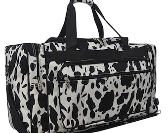 May include: A rectangular duffel bag featuring a black and white cow print pattern. The bag includes a large main compartment with a zipper closure, a front zippered pocket, two top handles, and black trim along the edges and zippers.