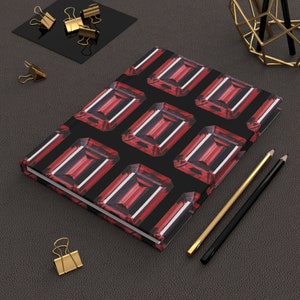 Può includere: Un taccuino nero con un motivo di pietre preziose rosse sulla copertina. Il taccuino è appoggiato su una superficie grigio scuro con graffette dorate e matite.