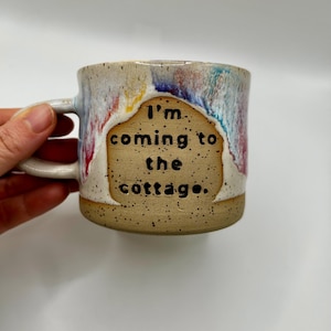以下が含まれることがあります： 手作りのセラミックマグカップ。ベージュの斑点のあるベースと、赤、青、黄色の色合いのカラフルなドリップ釉薬が施されています。「I'm coming to the cottage」の文字が黒い文字でタン色の背景に書かれています。