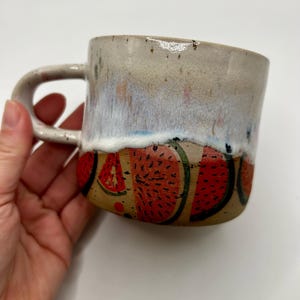 May include: A handmade ceramic mug with a watermelon design. The mug has a beige exterior with a white and blue glaze at the top. The bottom half features red watermelon slices with black seeds. The mug is held by a hand.