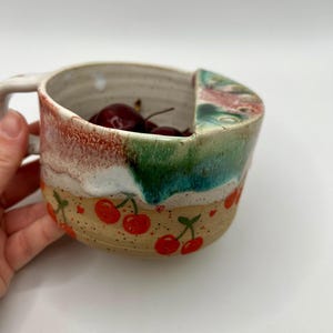 May include: A handmade ceramic mug with a unique design. The mug features a handle and a colorful exterior with red cherries, green, white, and red accents. The mug is filled with cherries.