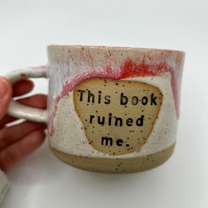 May include: A ceramic mug with a cream base and a red and white glaze. The mug features the text "This book ruined me" in black lettering on a tan patch. The mug has a curved handle. The mug is handmade.