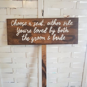 May include: A wooden sign with the text "Choose a seat, either side. You're loved by both the groom & bride." The sign is mounted on a wooden post.