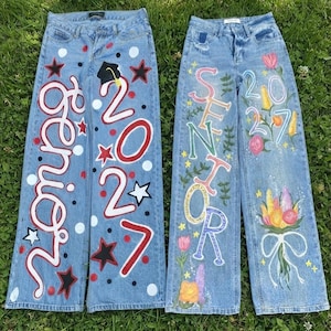 May include: Two pairs of light blue denim jeans featuring hand-painted designs. The left pair displays the word Senior, the year 2027, a graduation cap, and stars. The right pair features the word Senior, the year 2027, floral motifs, and a bouquet.