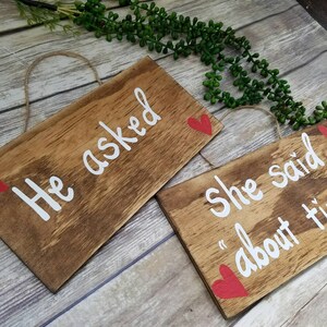 May include: Two wooden signs with red hearts and white lettering. The first sign says "He asked" and the second sign says "She said "about time"".