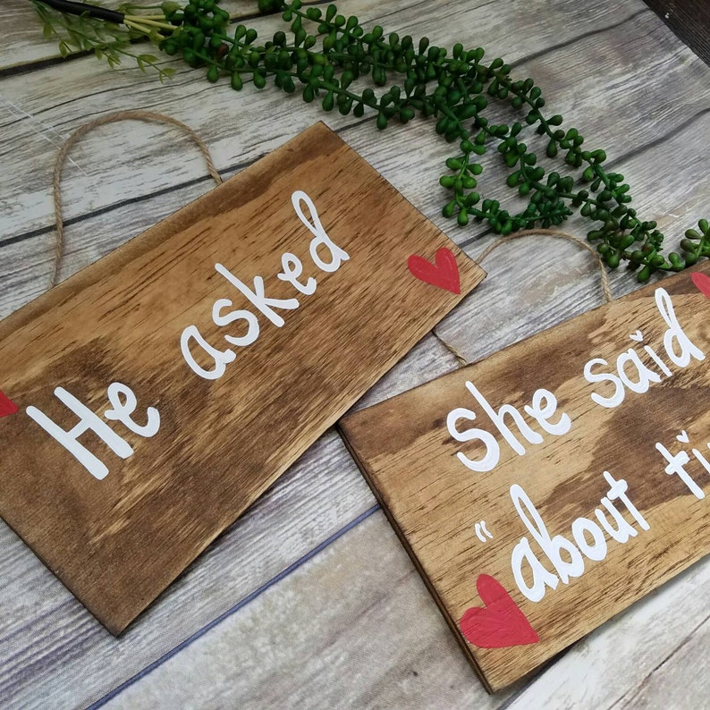 May include: Two wooden signs with red hearts and white lettering. The first sign says "He asked" and the second sign says "She said "about time"".