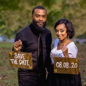 May include: A couple holding wooden signs with the text "Save the Date" and "08.08.20". The man is wearing a black shirt and the woman is wearing a white shirt with the word "Bride" on it.