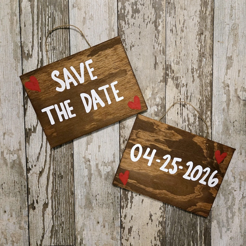 May include: Two wooden signs with white text and red hearts. The top sign says "Save the Date" and the bottom sign says "04-25-2026".