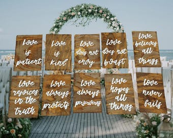 May include: A set of ten rectangular wooden signs with natural wood grain, each featuring white raised script lettering. The signs display the phrases: Love is patient, Love is kind, Love does not envy, Love does not boast, Love believes all things, Love rejoices in the truth, Love always protects, Love always perseveres, Love hopes all things, and Love never fails.