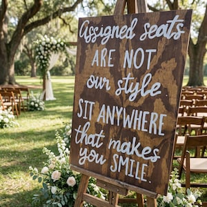 May include: A rectangular wooden sign with a dark brown stained finish and visible wood grain. The sign features white hand-lettered text that reads: Assigned seats ARE NOT our style SIT ANYWHERE that makes you SMILE. The sign is mounted on a wooden easel.