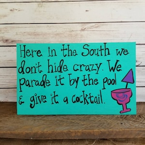 May include: A turquoise wooden sign with black text that reads "Here in the South we don't hide crazy. We parade it by the pool & give it a cocktail." A purple and pink cocktail glass with a purple umbrella is painted on the right side of the sign.