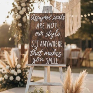 May include: A rectangular dark brown wooden sign displayed on a light-colored wooden easel. The sign features white hand-lettered text that reads: Assigned seats ARE NOT our style. SIT ANYWHERE that makes you SMILE.