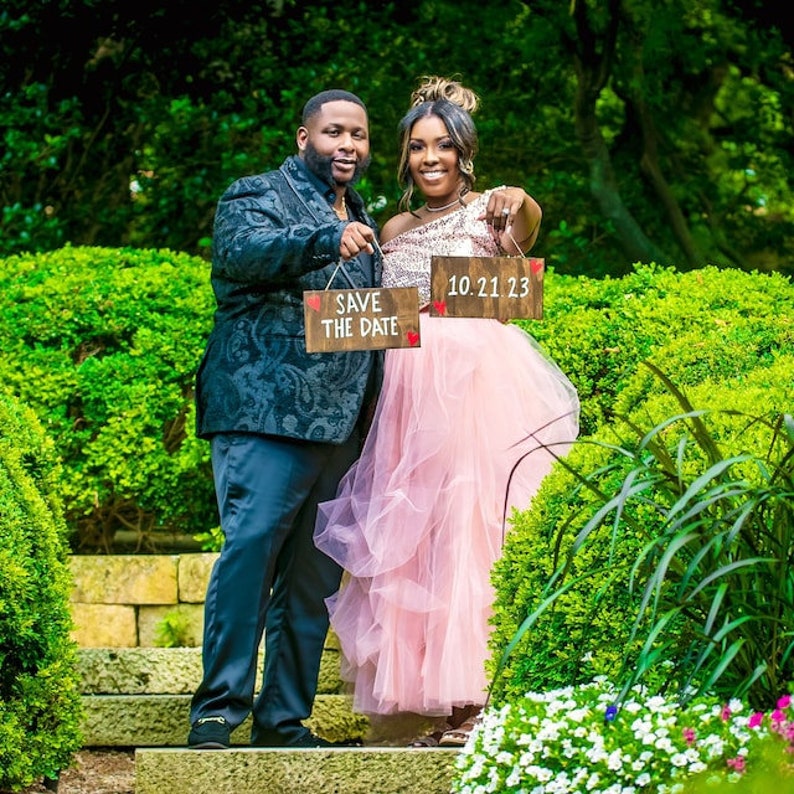May include: A couple pose for a photograph holding two wooden signs. The sign on the left says "Save the Date" with a red heart. The sign on the right says "10.21.23". The woman is wearing a pink tulle skirt and a gold top. The man is wearing a black suit jacket with a patterned design and black trousers.