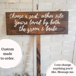 May include: Wooden sign with white text that reads "Choose a seat, either side. You're loved by both the groom & bride." The sign is on a wooden post and is in front of a white brick wall. The text "Custom made to order." is written in white on a circle in the lower left corner of the image. The text "I can change anything you'd like. Message me." is written in white on a circle in the lower right corner of the image.