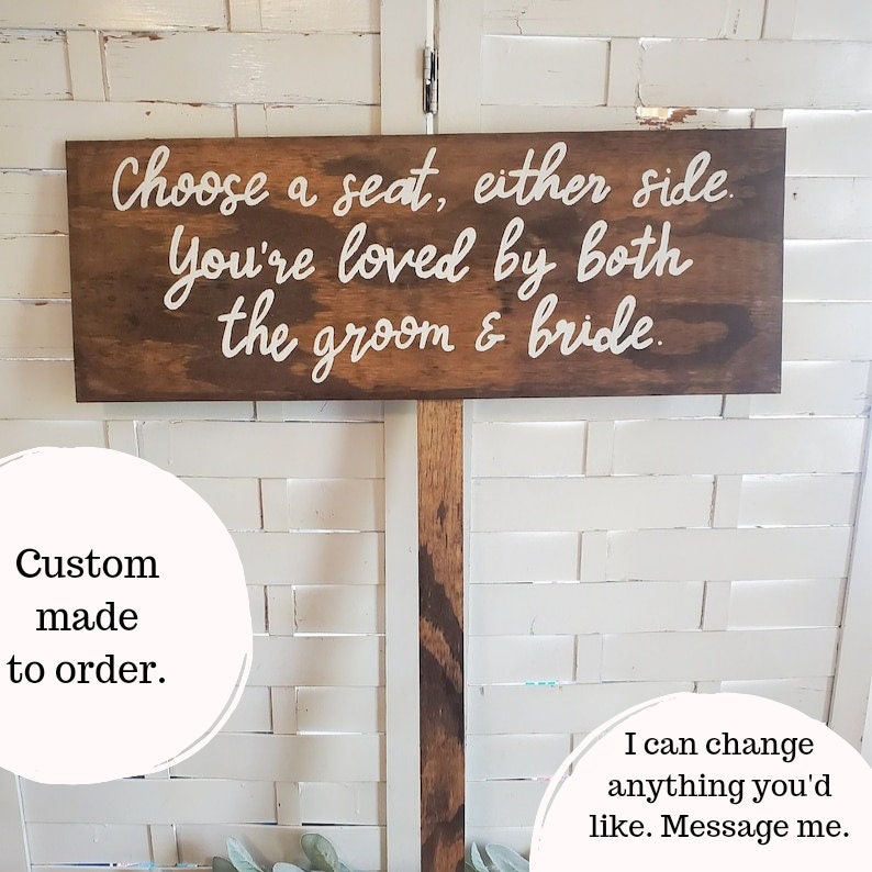 May include: Wooden sign with white text that reads "Choose a seat, either side. You're loved by both the groom & bride." The sign is on a wooden post and is in front of a white brick wall. The text "Custom made to order." is written in white on a circle in the lower left corner of the image. The text "I can change anything you'd like. Message me." is written in white on a circle in the lower right corner of the image.
