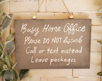 May include: A brown wooden sign with white text that reads "Busy Home Office Please DO NOT disturb Call or text instead Leave packages". The sign is hanging from a string with two wooden beads.