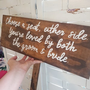 May include: A wooden sign with white lettering that reads "Choose a seat, either side. You're loved by both the groom & bride."