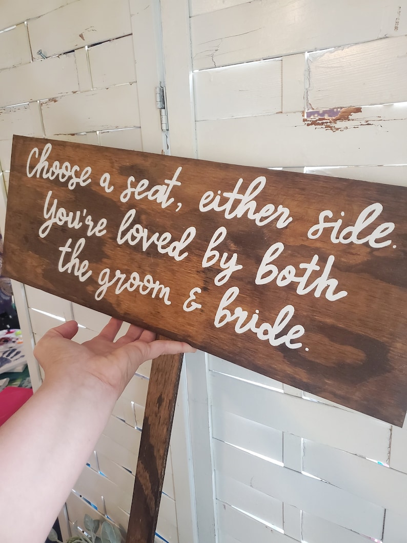 May include: A wooden sign with white lettering that reads "Choose a seat, either side. You're loved by both the groom & bride."