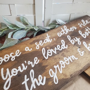May include: A wooden sign with white painted lettering that reads "Choose a seat, either side you're loved by both the groom & b".