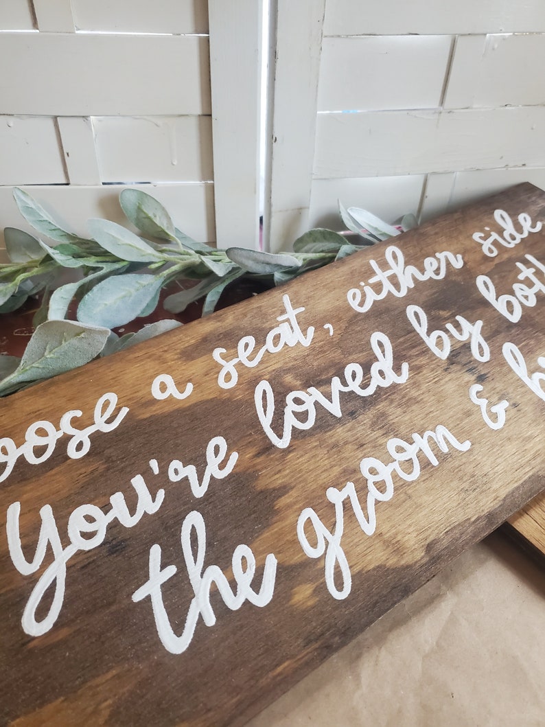 May include: A wooden sign with white painted lettering that reads "Choose a seat, either side you're loved by both the groom & b".