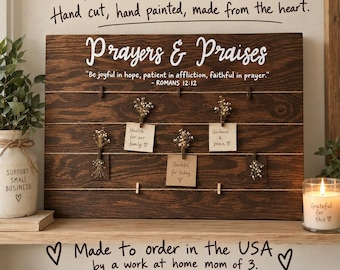 May include: A rectangular dark brown wooden board with horizontal twine lines and wooden clips. The text reads Prayers & Praises and Be joyful in hope, patient in affliction, faithful in prayer. Romans 12:12. Small paper notes with handwritten text are attached.