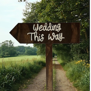 May include: A dark brown wooden arrow-shaped sign mounted on a vertical post. The sign features the white hand-lettered text Wedding This Way. The arrow points to the left, indicating a direction, with a visible wood grain texture throughout.
