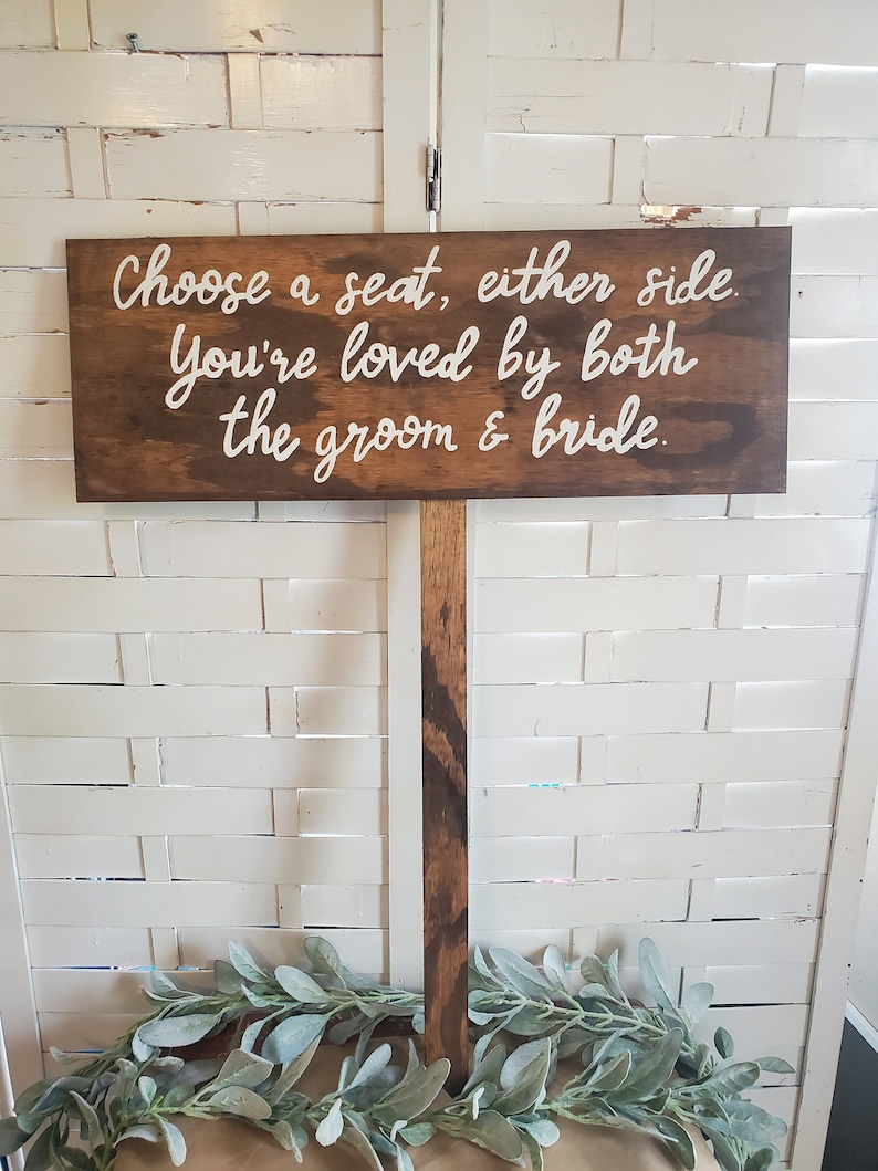 May include: A wooden sign with the text "Choose a seat, either side. You're loved by both the groom & bride." The sign is mounted on a wooden post.