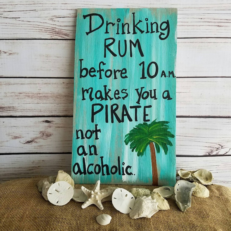 Pu&ograve; includere: Un cartello in legno blu turchese con strisce bianche dipinte e un motivo di palma. Il cartello recita "Drinking Rum before 10 a.m. makes you a Pirate not an alcoholic."