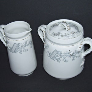 May include: A white porcelain creamer and sugar bowl set with a gray floral design and gold accents. The handles are rope-like and the sugar bowl has a lid.