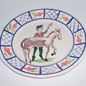 May include: A white ceramic plate with a blue and white checkered border. The center of the plate features a hand-painted illustration of a person riding a horse. The person is wearing a red shirt and green pants. The horse is brown and white.