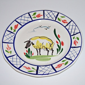 May include: A white ceramic plate with a blue and white checkered border. The center of the plate features a hand-painted illustration of a yellow sheep with black markings, surrounded by green and red flowers.