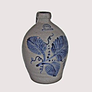 May include: A gray stoneware jug with a handle and a blue floral design. The jug features a blue leaf and vine pattern. The year "1996" is printed on the jug. The jug has a rounded body and a narrow neck.